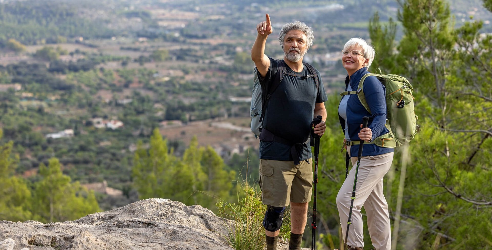 Ein älteres Paar steht mit Wanderstöcken auf einem Felsen und genießt den weiten Blick über eine grüne Hügellandschaft. Der Mann trägt eine GenuTrain OA Knieorthese zur Unterstützung bei Gonarthrose.