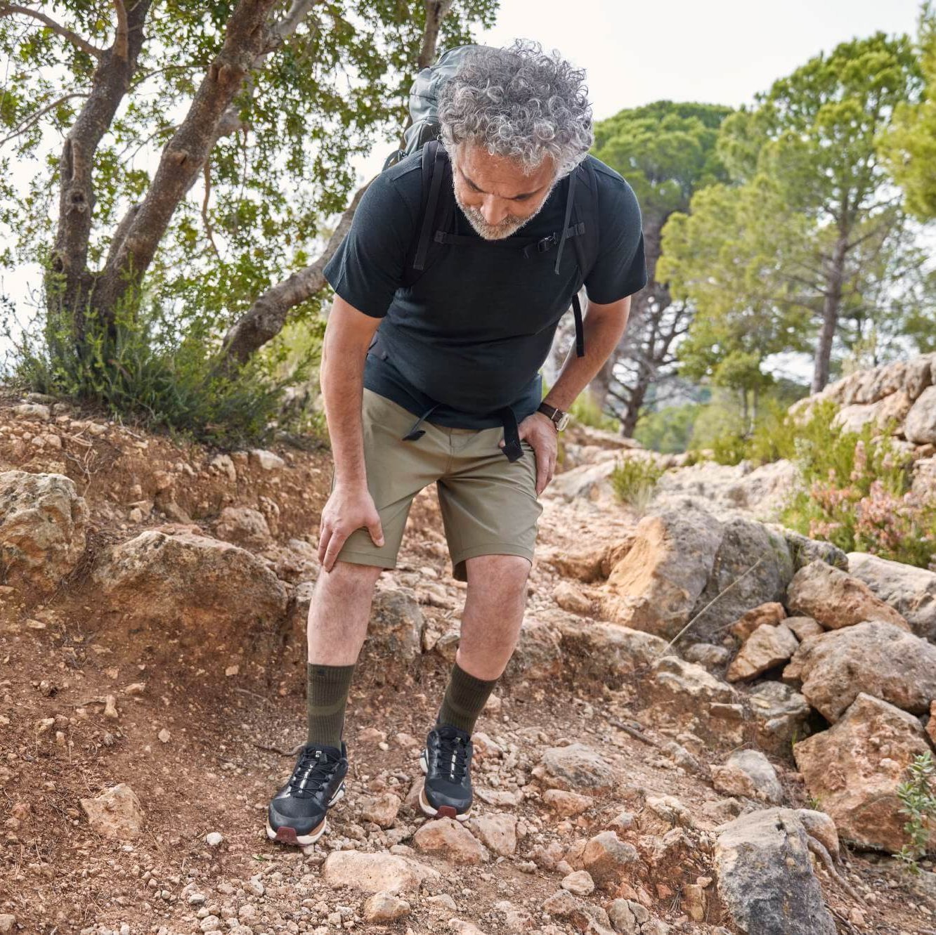 Ein älterer Mann mit grauen Locken und einem Rucksack hält auf einem steinigen Wanderweg inne und fasst sich ans schmerzende Knie. Er trägt ein schwarzes T-Shirt, beige Shorts und Wanderschuhe.
