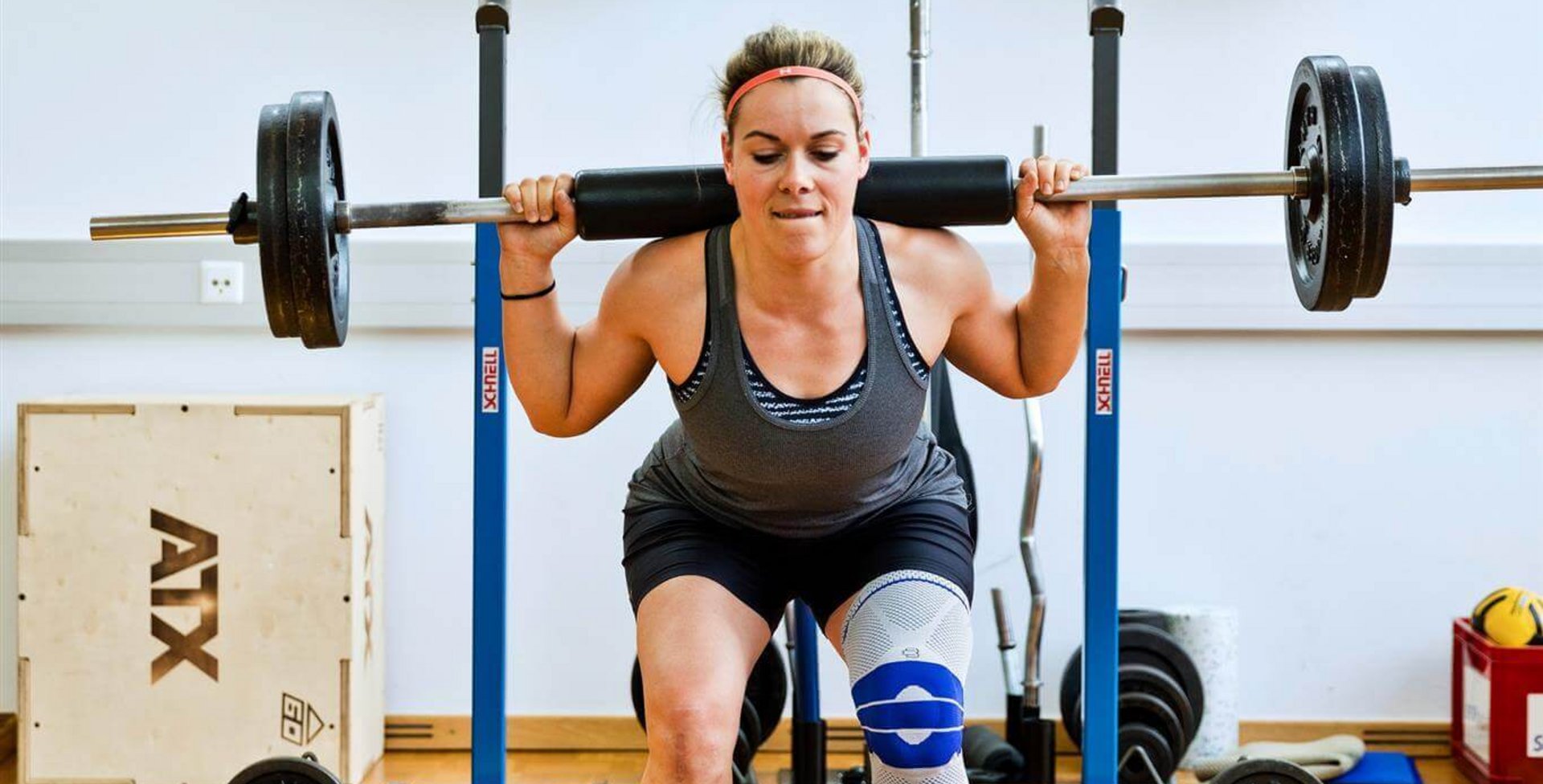 Andrea Limbacher stemmt Gewichte in einem Fitnessstudio. Sie trägt eine Bauerfeind GenuTrain Kniebandage am linken Knie, welche das Knie entlastet, stabilisiert und aktiviert.