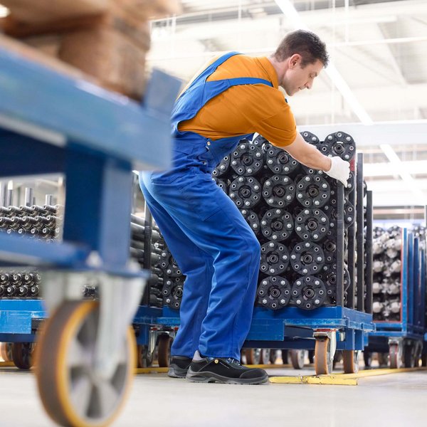 Das Bild zeigt einen jungen Mann in Arbeitskleidung, der einen Lastenwagen in einer Produktionshalle schiebt. Bei körperlicher Belastung ist es wichtig, die Gelenke immer wieder zu schonen und eintönige Bewegungen abwechslungsreich auszugleichen.