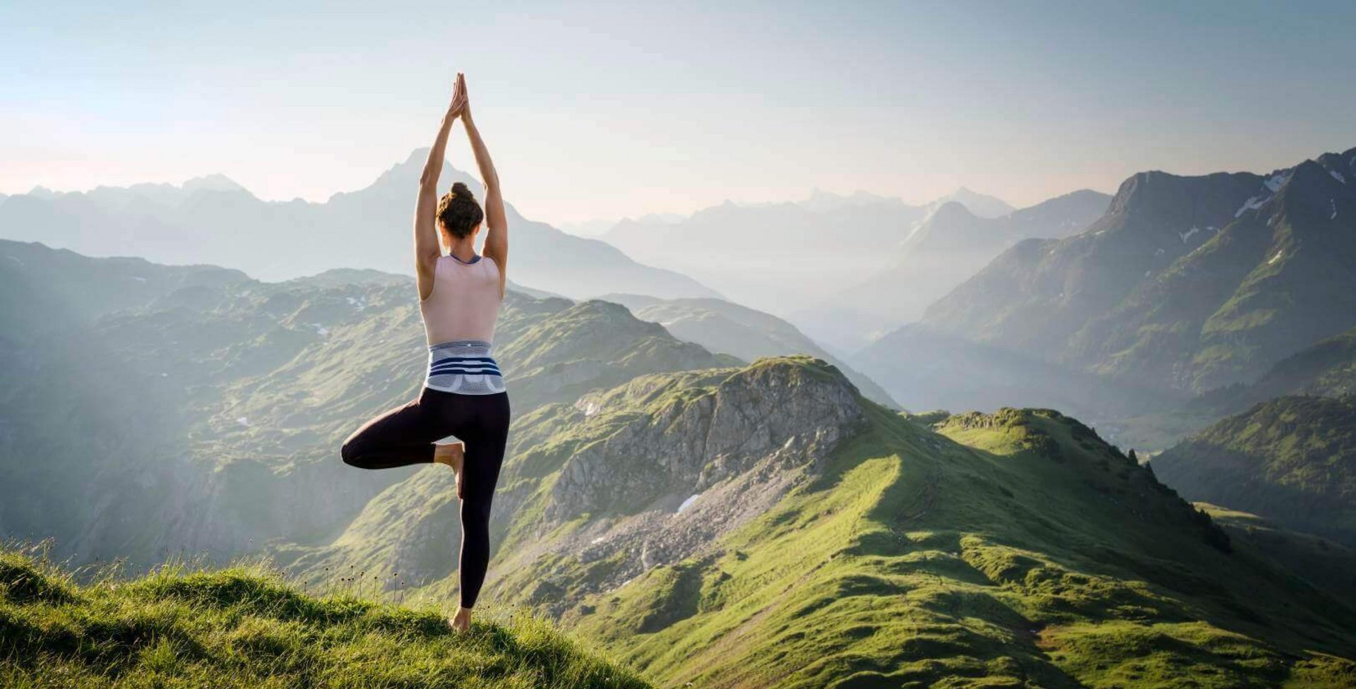 Das Bild zeigt eine Frau in Yogapose vor einem Bergpanorama. Yoga erweist sich als gelenkschonende Sportart. Zur Unterstützung ihres Rückens trägt die Frau eine LumboTrain Bandage.