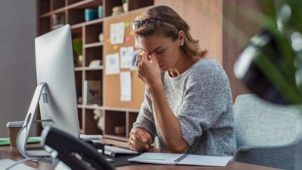 Das Bild zeigt eine gestresste Frau an ihrem Büroarbeitsplatz. Sie legt sich die Hand an die Stirn. Bewegungsmangel, z.B. bei der Arbeit, kann sich auch auf die Psyche auswirken.