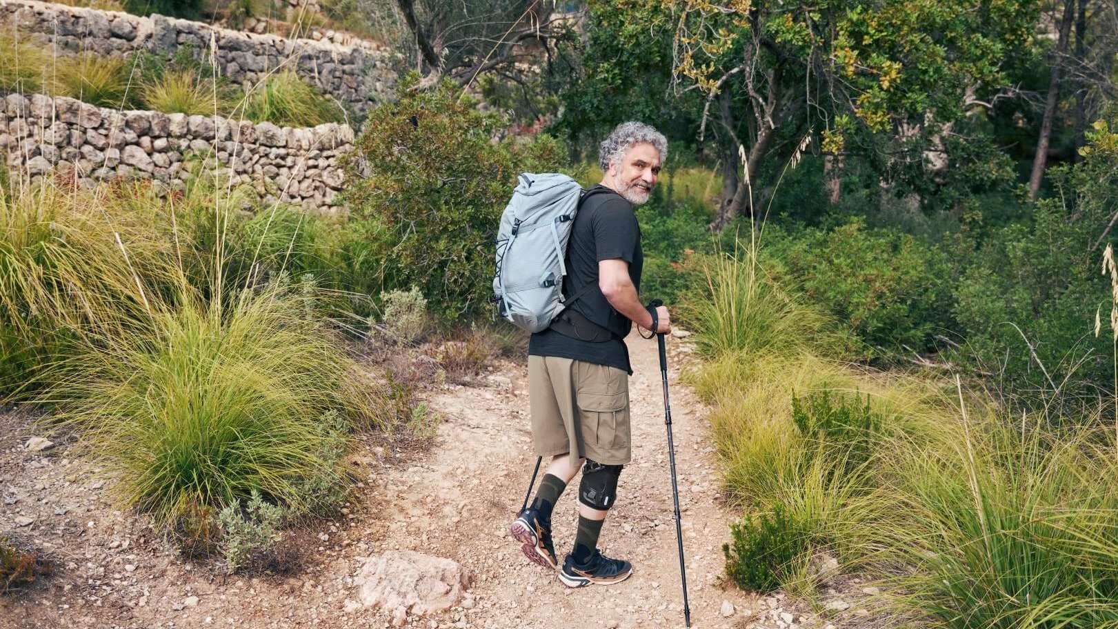 in älterer Mann mit grauen Locken wandert mit einem Trekkingrucksack und Wanderstöcken auf einem schmalen Naturpfad durch eine grüne Hügellandschaft. Er trägt eine GenuTrain OA Knieorthese zur Unterstützung bei Gonarthrose.