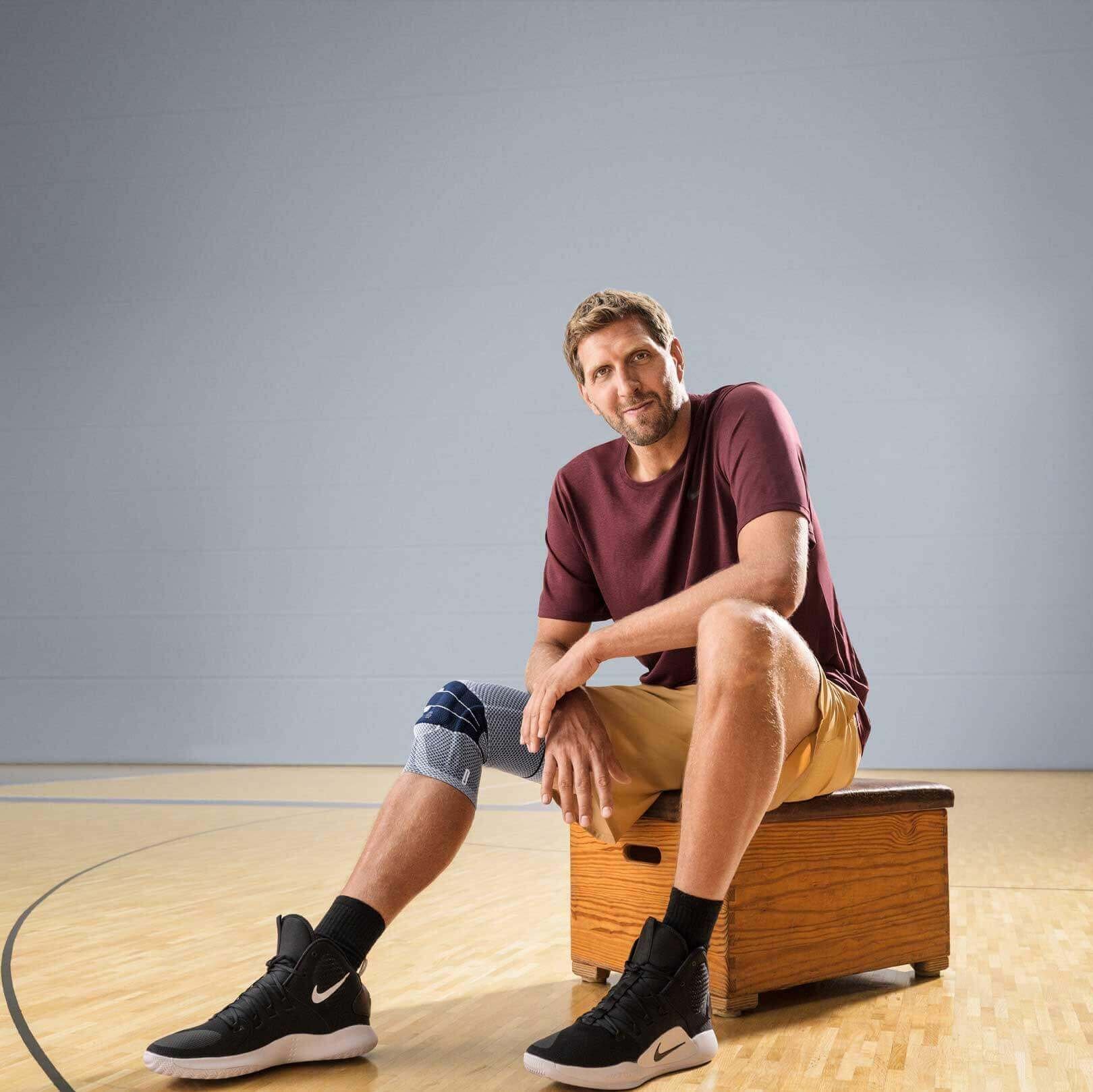 Dir Nowitzki sitzt auf einem Hocker in einer Sporthalle. Er trägt eine GenuTrain Kniebandage von Bauerfeind am rechten Knie. Er ist weltweiter Botschafter von Bauerfeind.