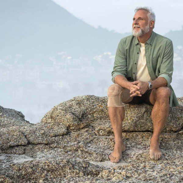 Das Bild zeigt einen älteren Mann, der an der Küste sitzt und den Ausblick genießt. Am Knie hat er eine GenuTrain Bandage zur Linderung von arthrosebedingten Schmerzen.