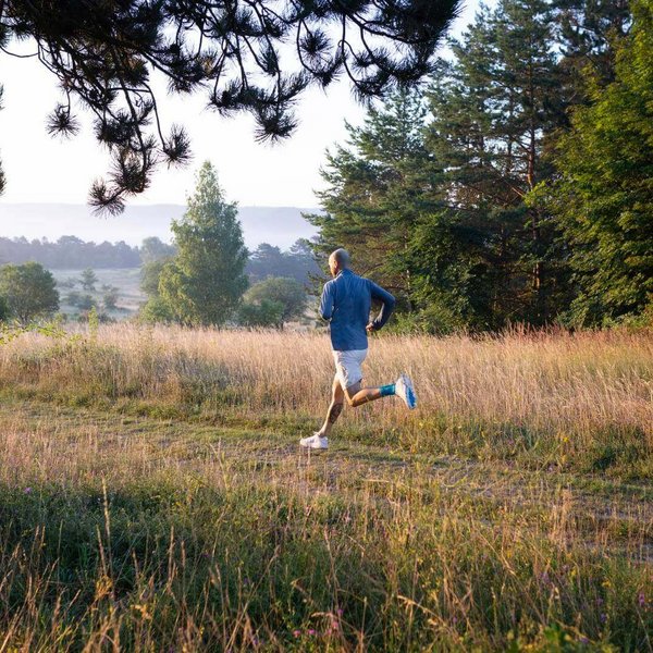 Abgebildet ist ein Läufer, der über eine Waldwiese rennt. Zur Unterstützung gegen Fersenschmerzen trägt er eine Bandage von Bauerfeind am Fußgelenk.