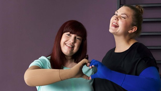 Auf dem Bild stehen zwei lächelnde Frauen nebeneinander und formen mit ihren Händen ein Herz. Beide tragen eine VenoTrain curaflow Armversorgung von Bauerfeind, die zur gezielten Ödemtherapie verwendet wird.