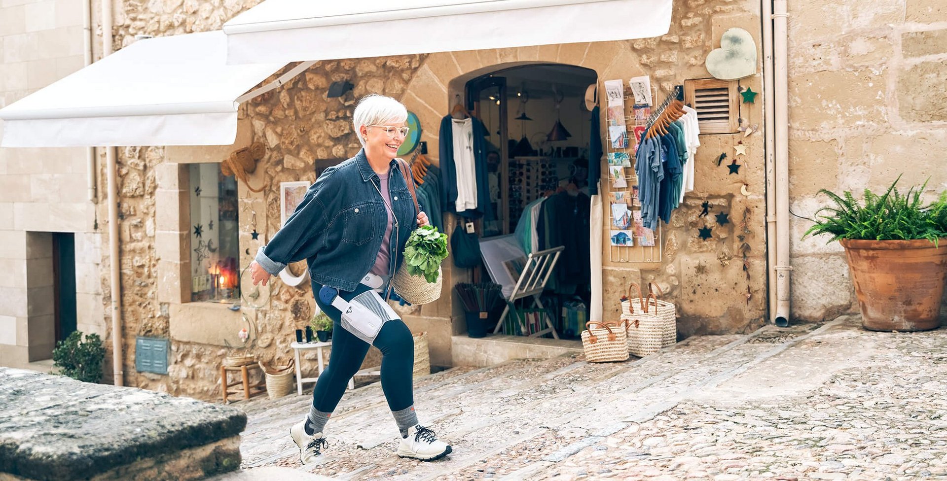 Eine ältere Frau mit kurzen weissen Haaren und Brille läuft eine gepflasterte Gasse hinauf. Sie trägt eine Jeansjacke, dunkle Leggings mit medizinischen Geräten daran und hält einen Korb mit frischem Grünzeug. Im Hintergrund befindet sich ein Geschäft mit Kleidung und Postkarten, eingerahmt von einer Steinwand mit weissen Markisen. Neben dem Eingang stehen Körbe und ein grosser Blumentopf.