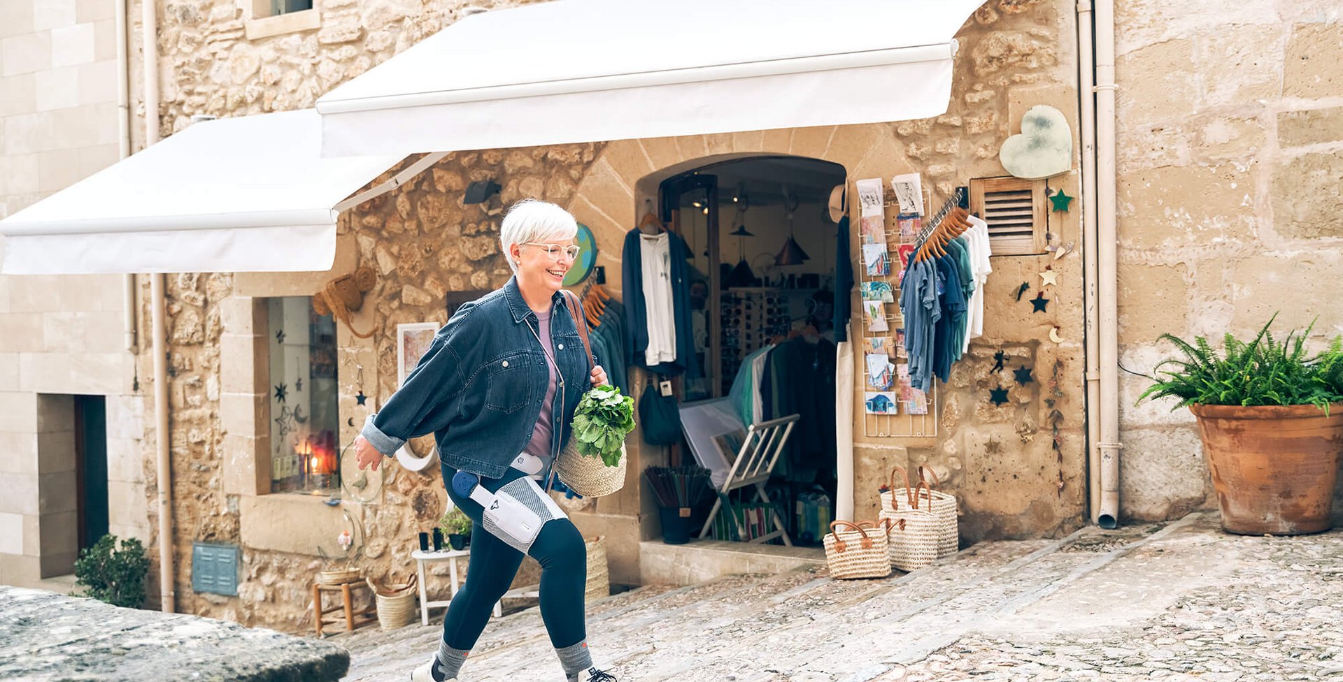 Eine ältere Frau mit kurzen weissen Haaren und Brille läuft eine gepflasterte Gasse hinauf. Sie trägt eine Jeansjacke, dunkle Leggings mit medizinischen Geräten daran und hält einen Korb mit frischem Grünzeug. Im Hintergrund befindet sich ein Geschäft mit Kleidung und Postkarten, eingerahmt von einer Steinwand mit weissen Markisen. Neben dem Eingang stehen Körbe und ein grosser Blumentopf.