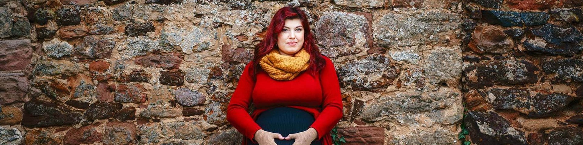 Abgebildet ist die schwangere Lipödem-Patientin Kathi Schanz, die an einer Steinmauer lehnt und ihre Hände auf ihren Bauch legt. Unterstützung bietet ihre VenoTrain curaflow Kompression.