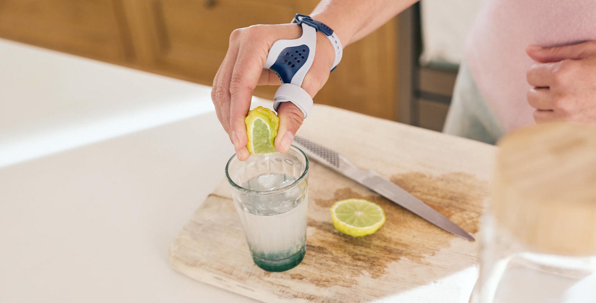 Nahaufnahme einer Hand, die mit der RhizoLoc OA Daumenorthese von Bauerfeind eine Zitrone über einem Glas ausdrückt, auf einem Schneidebrett mit einer angeschnittenen Zitrone und einem Messer im Hintergrund.