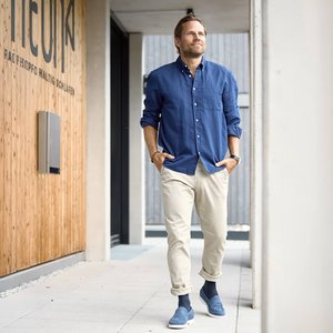 Mann in blauem Hemd und heller Hose geht mit den Händen in den Hosentaschen durch einen modernen Gebäudegang.