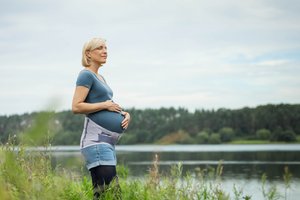 Schwangere Frau steht am Ufer eines Sees und hält ihren Babybauch, während sie in die Ferne blickt. Sie trägt die Spinova Mum Schwangerschaftsorthese und genießt die ruhige Naturkulisse.