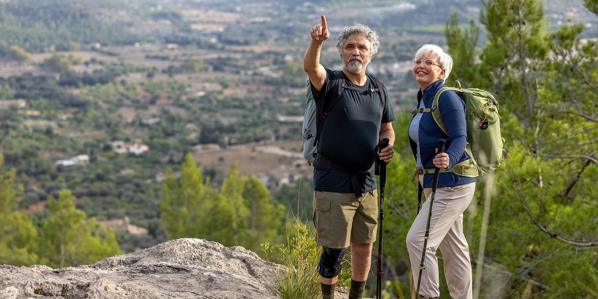 Ein älteres Paar steht mit Wanderstöcken auf einem Felsen und genießt den weiten Blick über eine grüne Hügellandschaft. Der Mann trägt eine GenuTrain OA Knieorthese zur Unterstützung bei Gonarthrose.