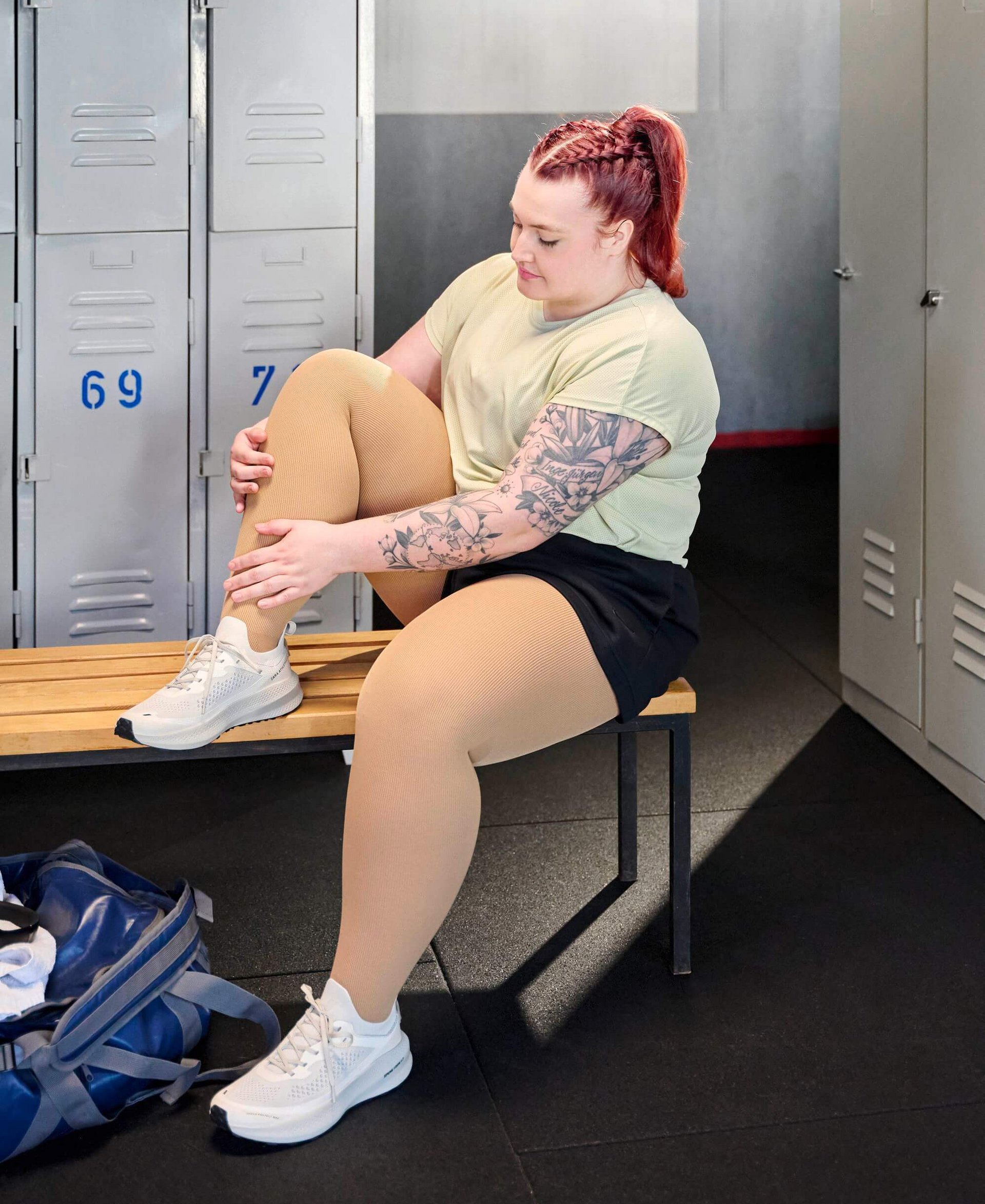Person sitzt auf einer Holzbank in einer Umkleidekabine und hält ihr angewinkeltes Bein, das mit cremefarbenen Kompressionsstrümpfen bekleidet ist. Sie trägt ein helles Sportshirt, schwarze Shorts und weisse Sportschuhe. Im Hintergrund graue Spinde mit blauen Nummern.