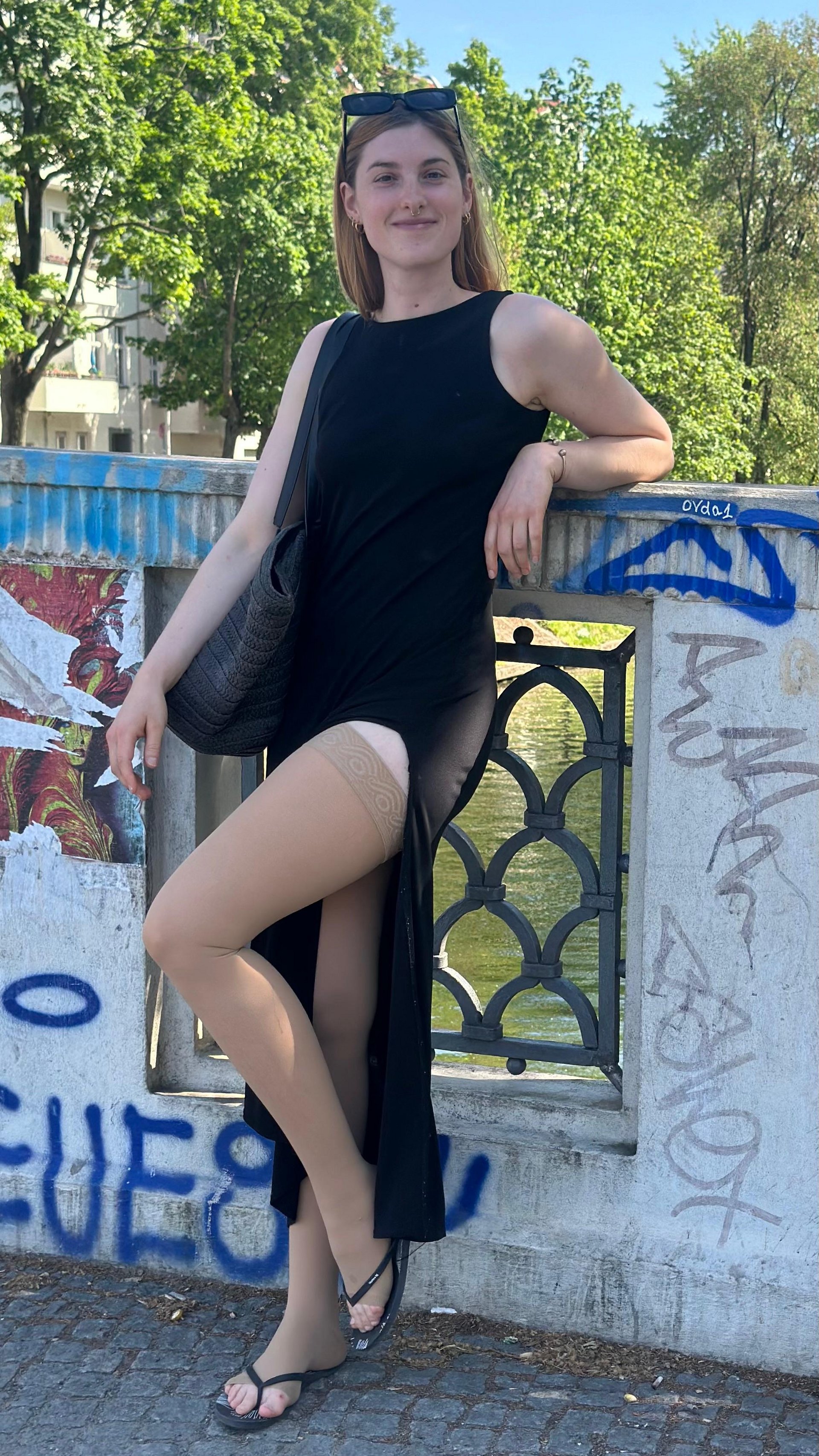 Eva Seemann trägt VenoTrain micro Kompressionsstrümpfe von Bauerfeind und lehnt lächelnd an einem Geländer in einer urbanen Umgebung. Sie trägt ein schwarzes Kleid und Sandalen.