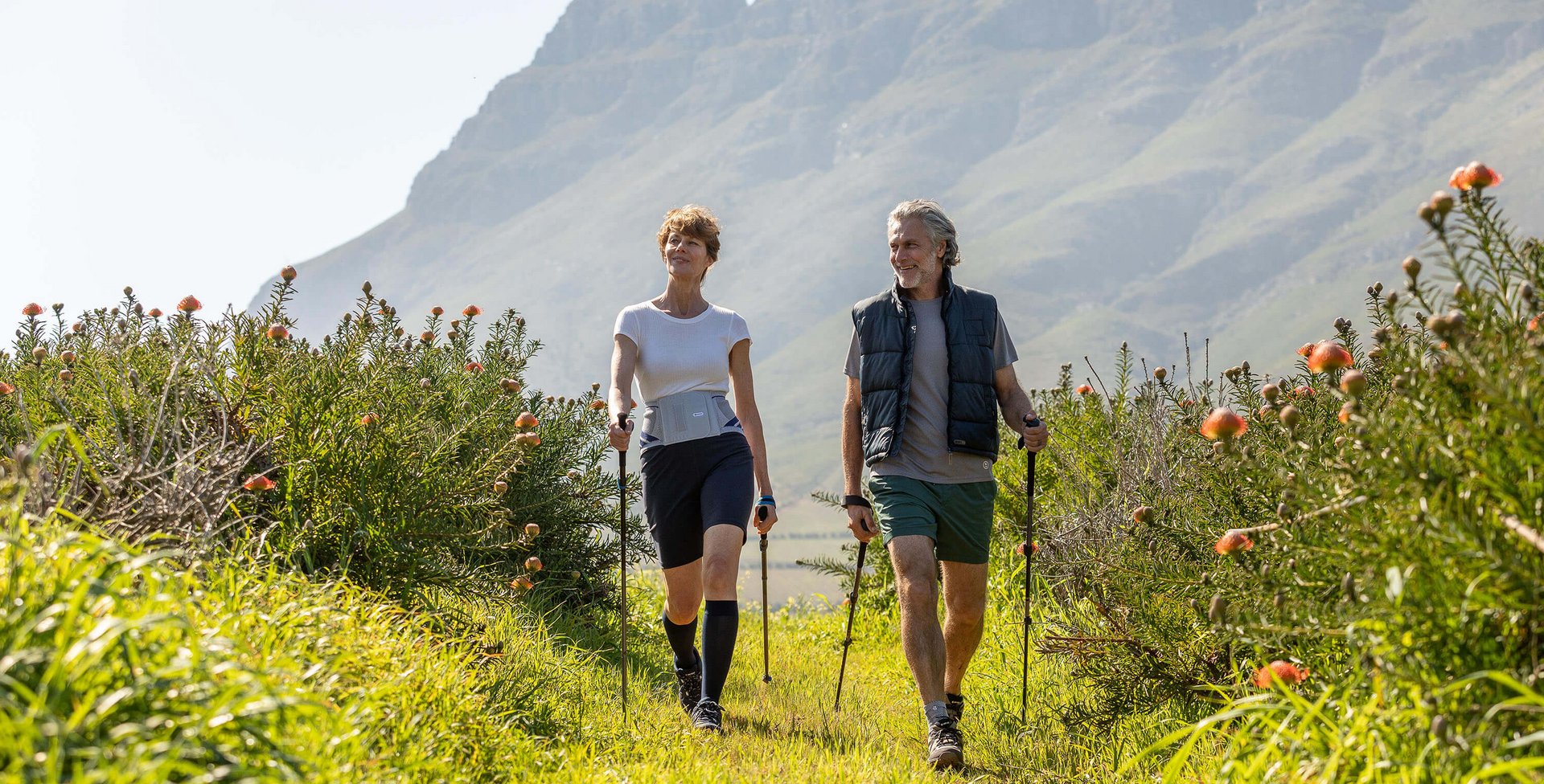 Auf dem Bild kann man sehen, wie ein älteres Paar in der Natur Nordic Walking betreibt. Sie tragen Train-Bandagen von Bauerfeind, um ihre Gelenke und den Rücken während der Bewegung zu entlasten.