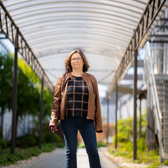 Katrin, eine Lymphödempatientin, steht selbstbewusst in einer überdachten Passage. Sie trägt eine braune Lederjacke und Jeans, das Licht fällt hell auf die Umgebung, während Pflanzen auf beiden Seiten des Weges für eine freundliche Atmosphäre sorgen.