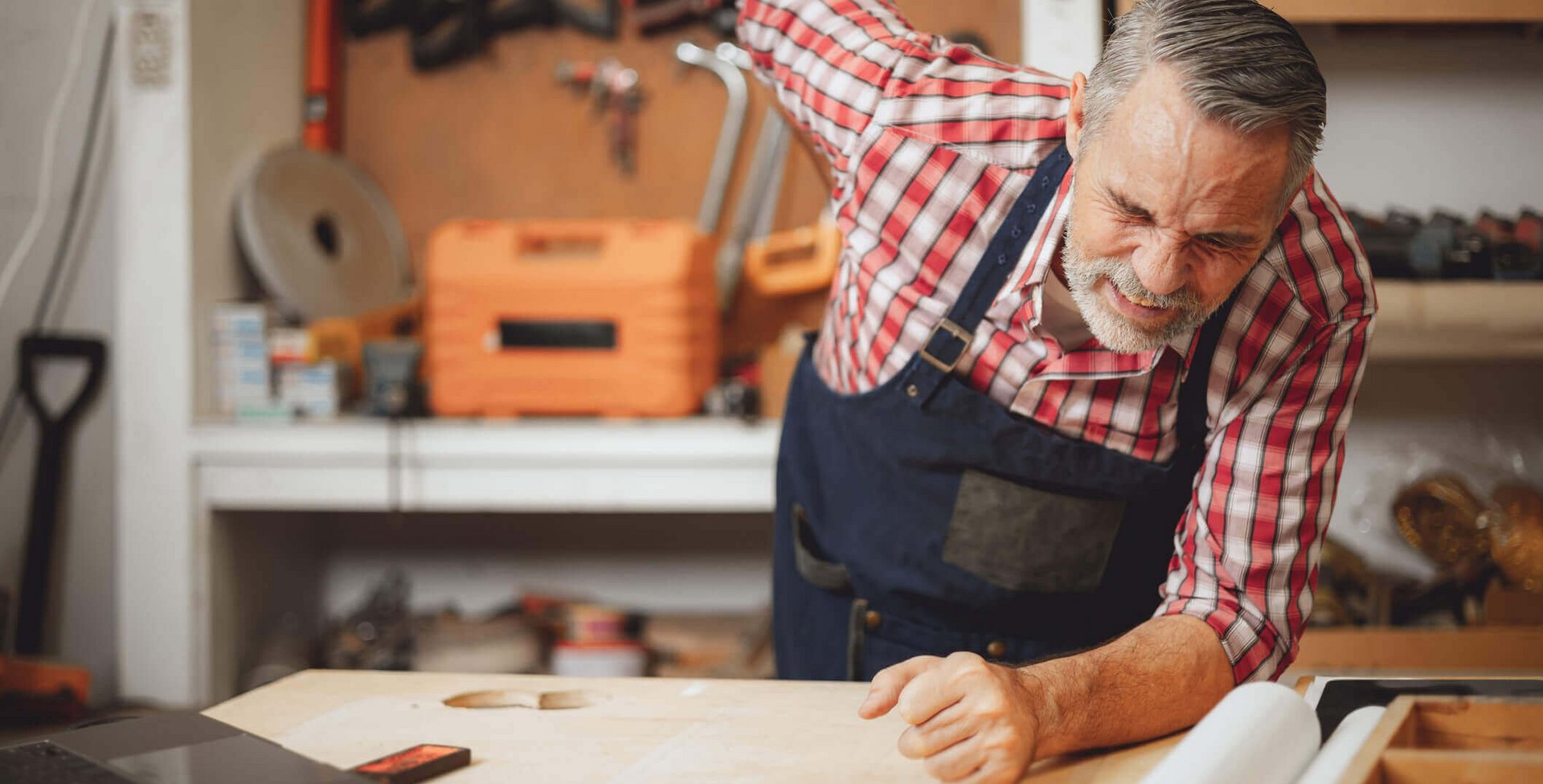 Abgebildet ist ein Handwerker in seiner Werkstatt, der sich mit schmerzverzehrtem Gesicht an die Hüfte fasst. Hüftschmerzen sollte man unmittelbar angehen, um chronischen Schmerzen vorzubeugen.