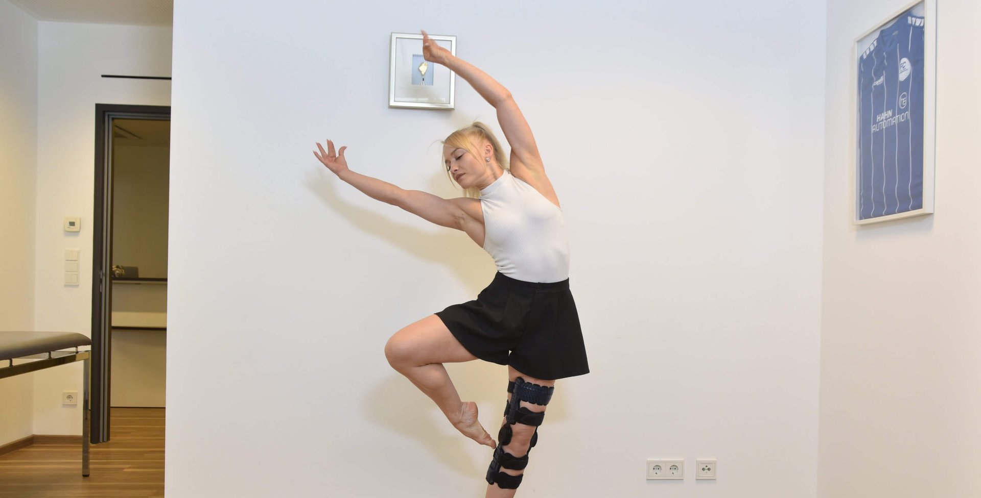 Anne-Maria Kot tanzt barfuß in einem Raum, trägt einen schwarzen Rock, ein weißes ärmelloses Oberteil und eine SecuTec Genu Flex Knieorthese am linken Bein.