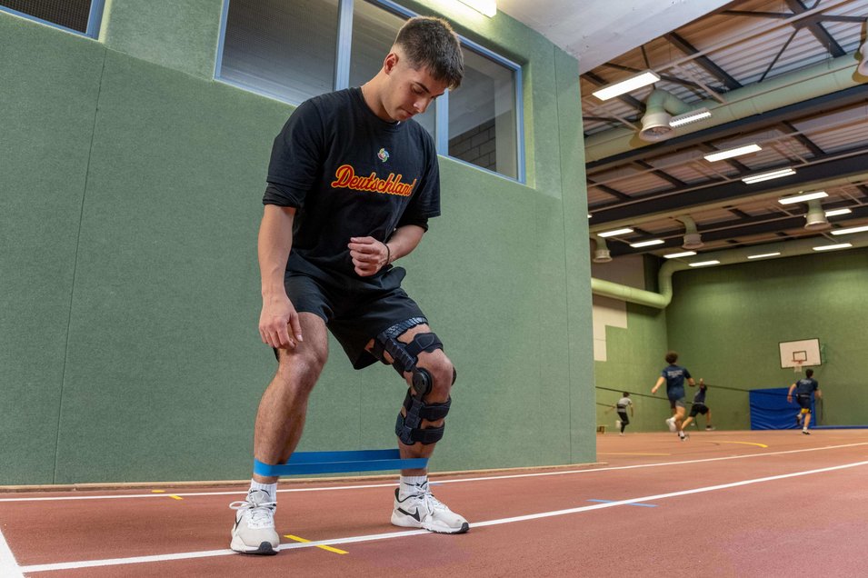 Simon Liedkte, professioneller Baseballspieler, führt Übungen in einer Turnhalle durch. Er trägt eine SecuTec Genu Flex Knieorthese zur Stabilisierung seines hinteren Kreuzbandrisses. Im Hintergrund sind weitere Spieler beim Training zu sehen.