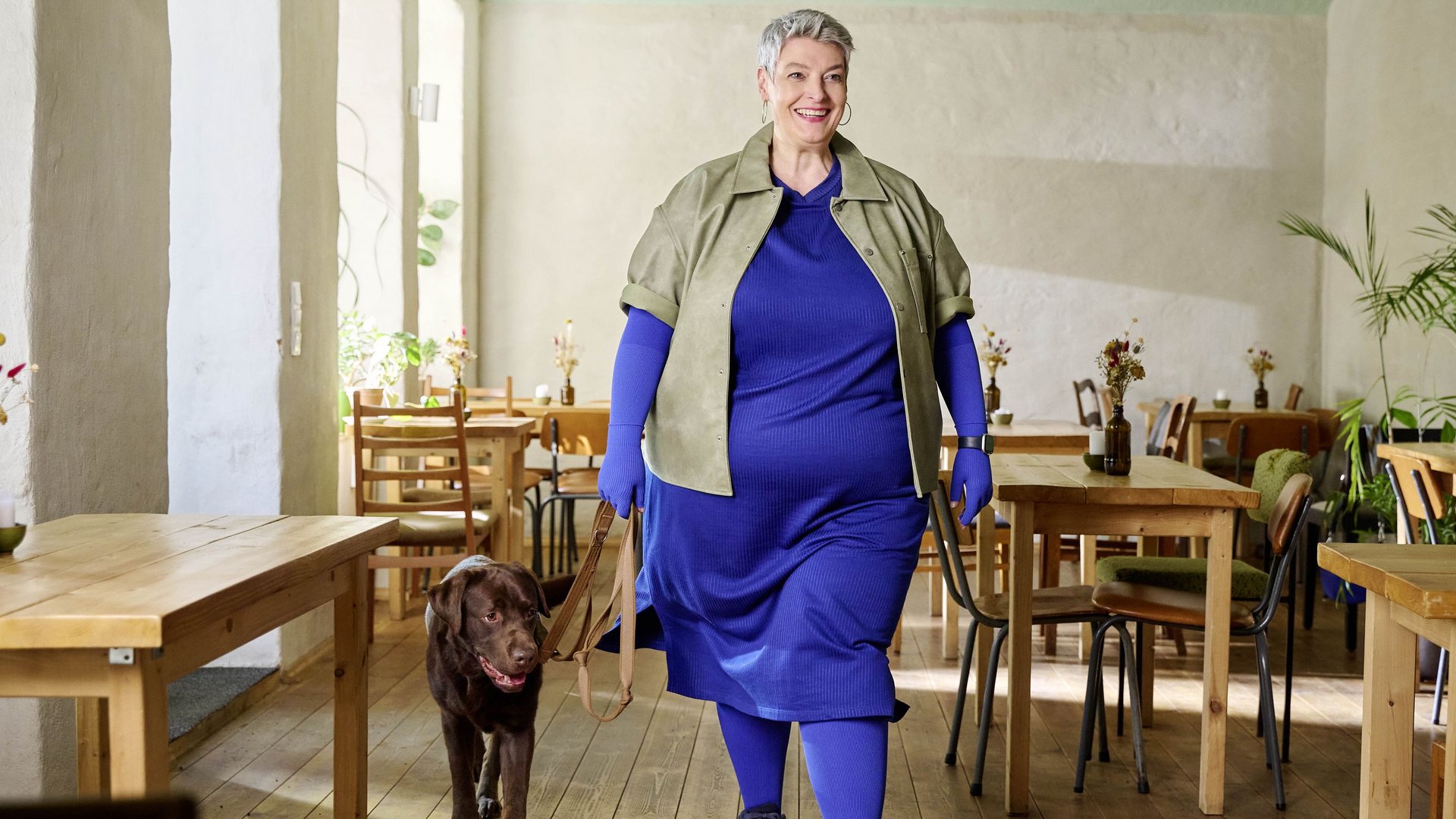 Frau in einem blauen Kleid, olivgrüner Jacke und blauen Kompressionshandschuhen, wie sie mit einem braunen Labrador durch ein helles Café mit Holztischen geht.