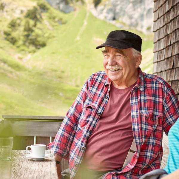 Ein Ehepaar sitzt an einem Tisch an einer Berghütte und macht eine Pause. Sie schauen beide freudig in die Ferne. Hallux Rigidus Einlagen von Bauerfeind schaffen Abhilfe, wenn die durch Arthrose beeinträchtigte Großzehe das Laufen zur Tortur macht.