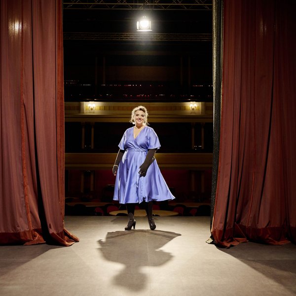 Eine Frau steht in einem eleganten, blauen Kleid auf einer Theaterbühne zwischen zwei roten Vorhängen, frontal beleuchtet von einem Scheinwerfer. Der Theatersaal im Hintergrund ist schwach beleuchtet.