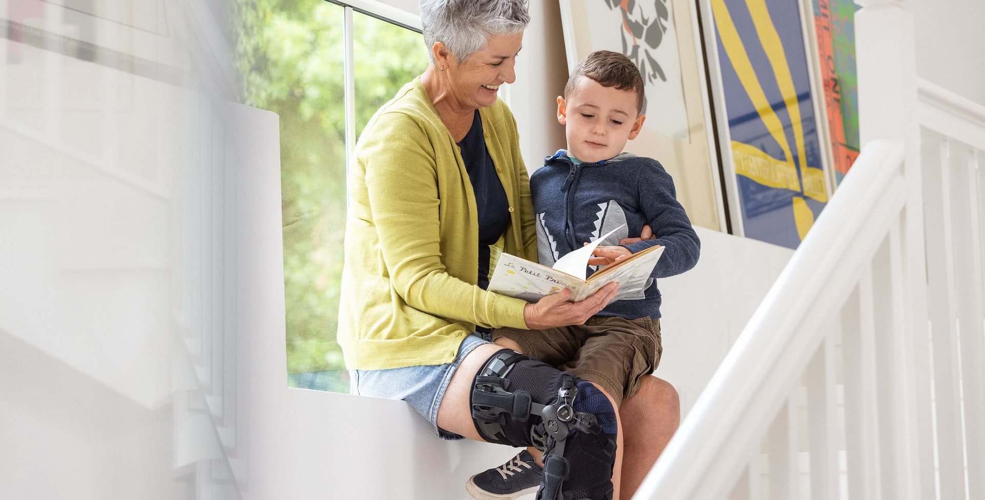 Arthrose im Knie: Was hilft? Eine ältere Frau sitzt auf einer Fensterbank und liest einem kleinen Jungen ein Buch vor. Sie trägt eine GenuTrain OA Knieorthese am rechten Bein. Im Hintergrund sind bunte gerahmte Bilder an der Wand zu sehen.