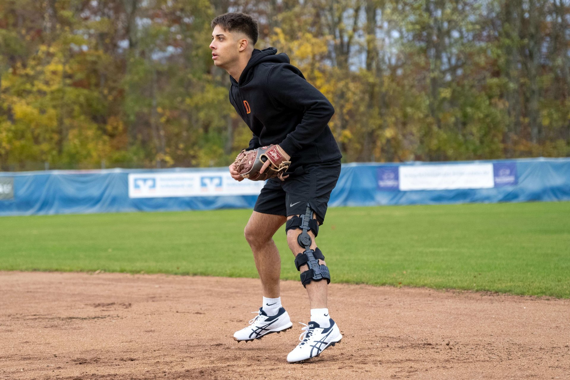 Simon Liedkte steht in der Feldverteidigungsposition auf einem Baseballplatz. Er trägt eine schwarze Kapuzenjacke, Shorts und die SecuTec Genu Flex Knieorthese am rechten Bein, um sein Knie nach einem hinteren Kreuzbandriss zu stabilisieren. 