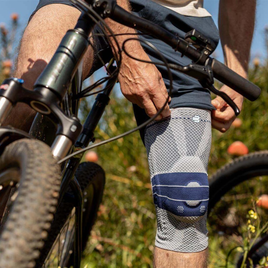 Der Bildausschnitt zeigt einen Fahrradfahrer, der anhält, um seine GenuTrain Kniebandage anzulegen. Diese Bandage bietet Unterstützung gegen Schmerzen während des Radfahrens.