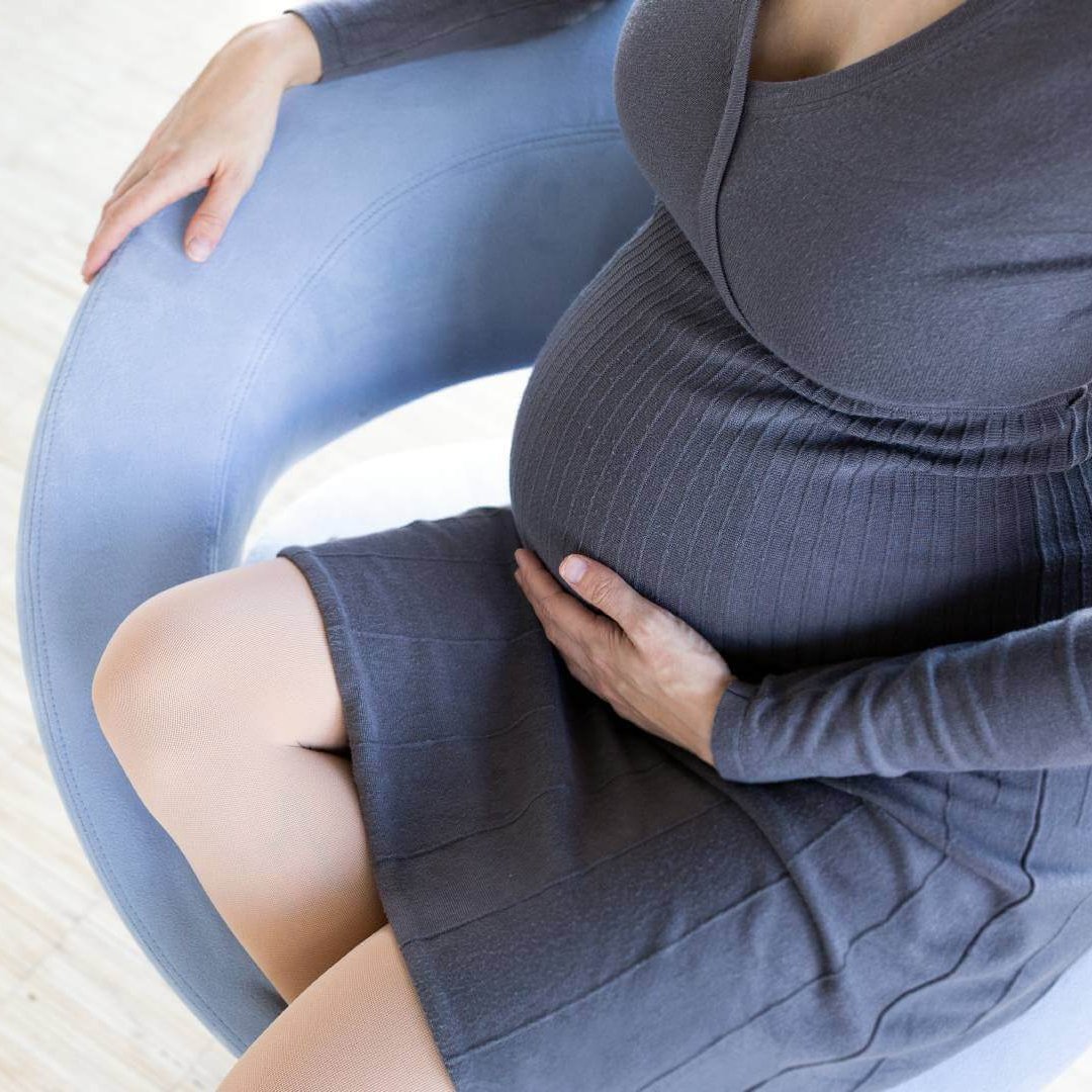 Medizinische Kompressionsstrümpfe in der Schwangerschaft Eine schwangere Frau im weißen Kleid steht in einem Hof und lächelt freundlich in die Kamera. Sie trägt einen Cardigan, welcher im Winde weht. Außerdem hat sie eine VenoTrain® micro Schwangerschaftsstrumpfhose von Bauerfeind an, welche die werdende Mutter optimal versorgt.