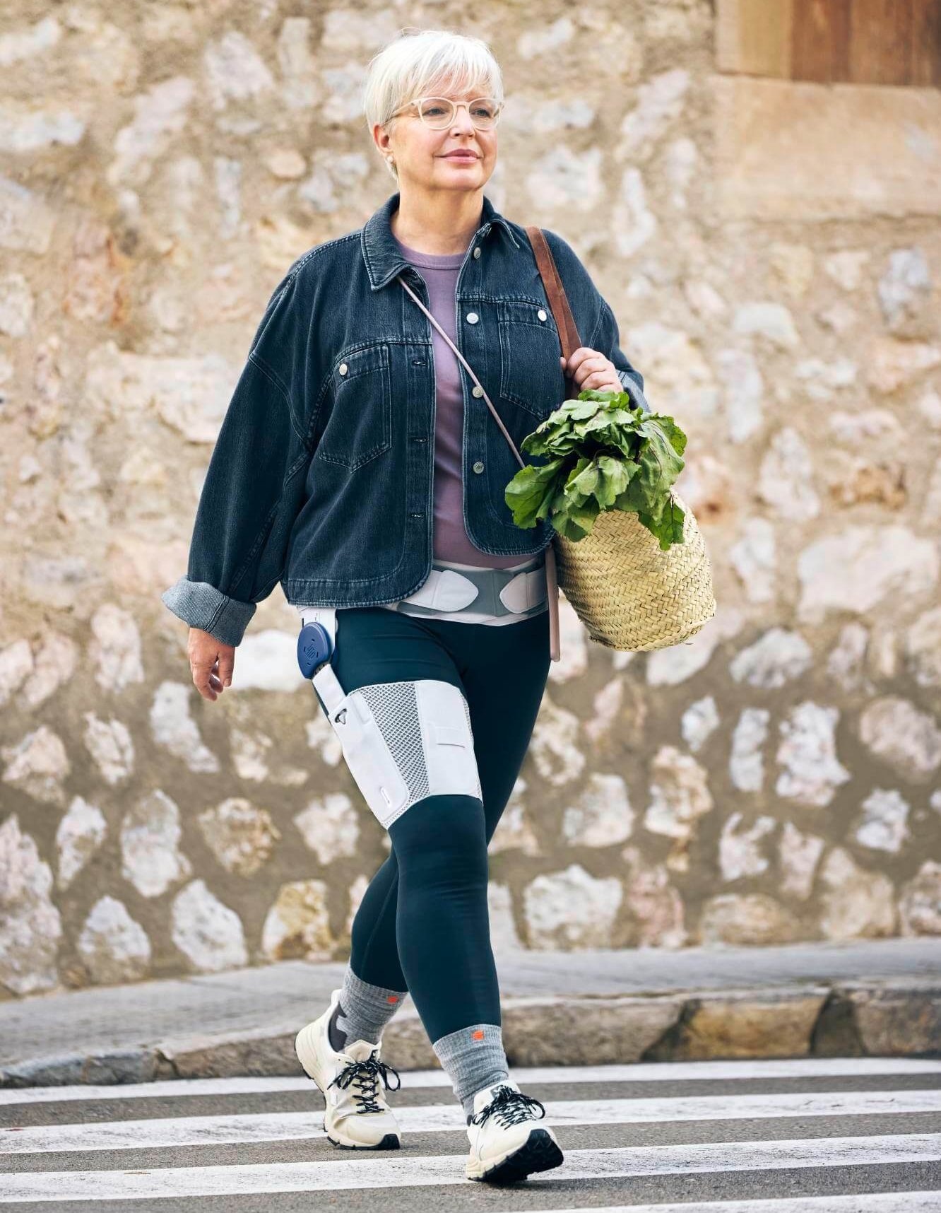 Eine ältere Frau mit kurzen weissen Haaren und Brille überquert einen Zebrastreifen. Sie trägt eine Jeansjacke, ein rosafarbenes Oberteil und dunkle Leggings, an denen medizinische Geräte befestigt sind. In ihrer Hand hält sie einen Korb mit frischem Grünzeug. Im Hintergrund ist eine Steinmauer zu sehen.