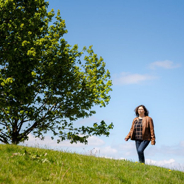 Katrin, eine Lymphödempatientin, spaziert über eine grüne Wiese unter einem klaren, blauen Himmel. Im Hintergrund steht ein großer, voller Baum, der eine sommerliche und ruhige Atmosphäre vermittelt. Sie trägt eine braune Lederjacke und Jeans.