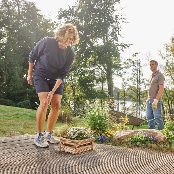 Eine Frau greift sich beim Gärtnern an ihr schmerzendes Knie. Im Hintergrund steht ein Mann mit Handschuhen und schaut zu. Der Garten ist von bunten Blumen und einer idyllischen Umgebung mit Bäumen und einem See geprägt.