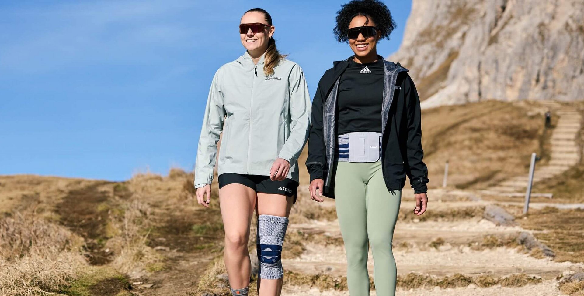 Laura Nolte und Deborah Levi wandern auf einem sonnigen Bergpfad mit felsigem Untergrund. Beide tragen Outdoor-Bekleidung und Sonnenbrillen; Laura trägt eine Kniebandage und Deborah eine Rückenbandage über ihrer Kleidung. Im Hintergrund sind ein trockener Grasbereich, ein steiler Pfad mit Stufen sowie felsige Bergformationen zu sehen.