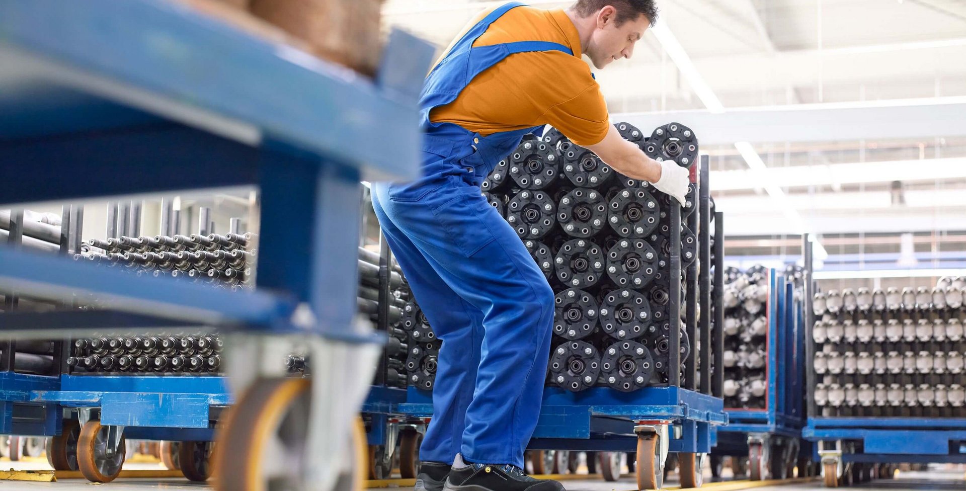 Das Bild zeigt einen jungen Mann in Arbeitskleidung, der einen Lastenwagen in einer Produktionshalle schiebt. Bei körperlicher Belastung ist es wichtig, die Gelenke immer wieder zu schonen und eintönige Bewegungen abwechslungsreich auszugleichen.