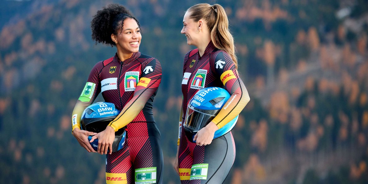 Zwei Bobfahrerinnen, Laura Nolte und Deborah Levi, in roten Rennanzuegen stehen nebeneinander im Freien vor einer Bergkulisse und blicken in die Kamera, beide tragen Sportbekleidung mit Logos und wirken selbstbewusst.