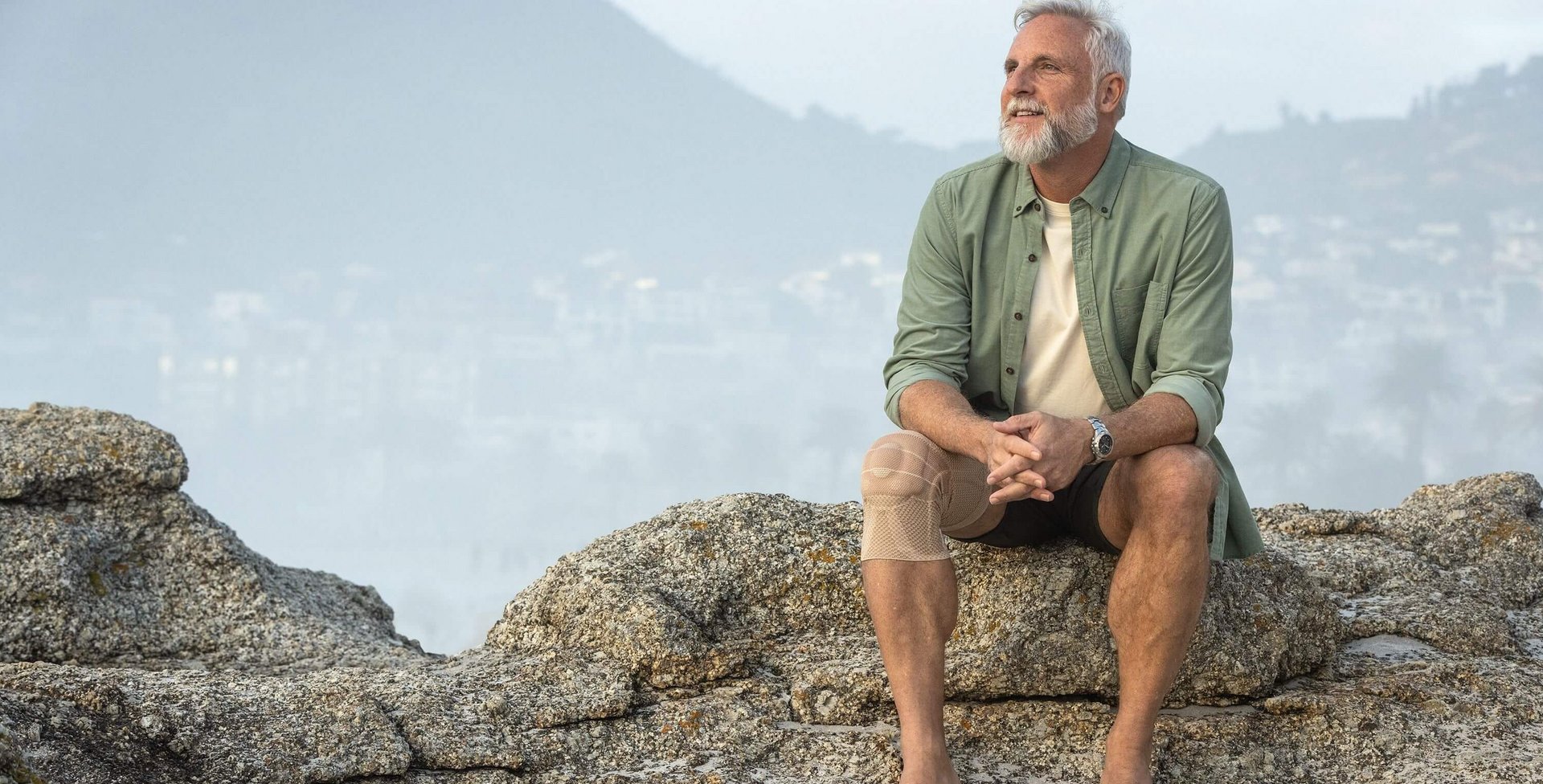 Das Bild zeigt einen älteren Mann, der an der Küste sitzt und den Ausblick genießt. Am Knie hat er eine GenuTrain Bandage zur Linderung von arthrosebedingten Schmerzen.