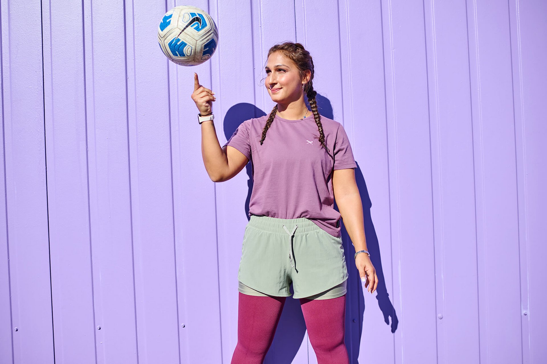 Junge Frau in sportlicher Kleidung steht vor einer lila Wand und balanciert einen Ball mit blauem Muster auf ihrem Finger. Sie trägt ein violettes T-Shirt, mintgrüne Shorts über bordeauxfarbenen Leggings und hat zwei geflochtene Zöpfe.