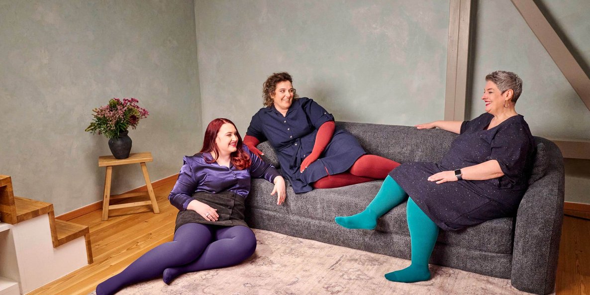 Drei Frauen sitzen und liegen entspannt auf einem dunkelgrauen Sofa in einem modern eingerichteten Wohnzimmer mit Holzfussboden. Sie tragen farbenfrohe Strumpfhosen – in Violett, Rot und Türkis – kombiniert mit dunklen Kleidern oder Röcken. Alle wirken fröhlich und unterhalten sich miteinander. Links steht ein kleiner Holztisch mit einer schwarzen Vase und einem Blumenstrauss.