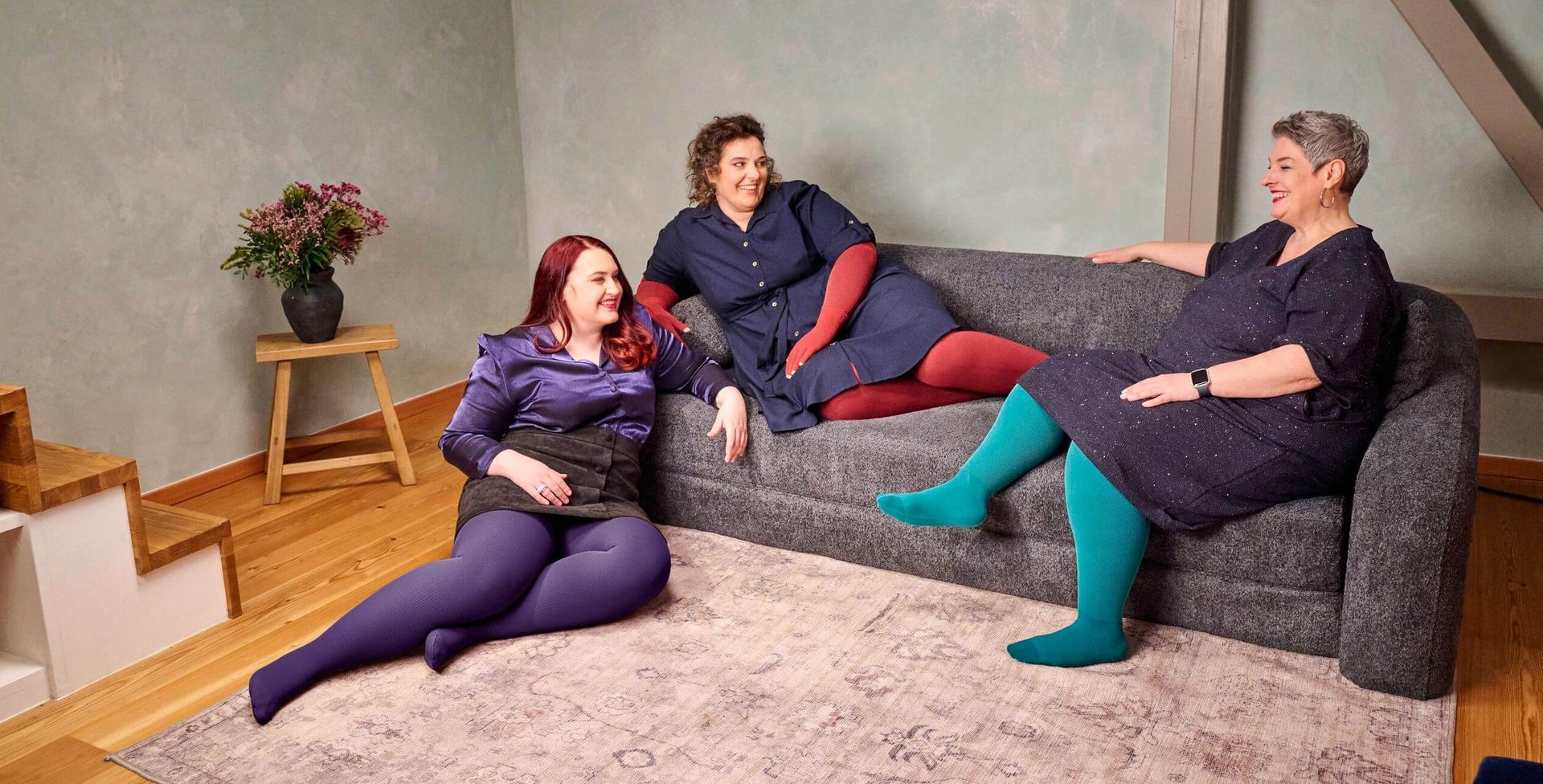 Drei Frauen sitzen und liegen entspannt auf einem dunkelgrauen Sofa in einem modern eingerichteten Wohnzimmer mit Holzfussboden. Sie tragen farbenfrohe Strumpfhosen – in Violett, Rot und Türkis – kombiniert mit dunklen Kleidern oder Röcken. Alle wirken fröhlich und unterhalten sich miteinander. Links steht ein kleiner Holztisch mit einer schwarzen Vase und einem Blumenstrauss.