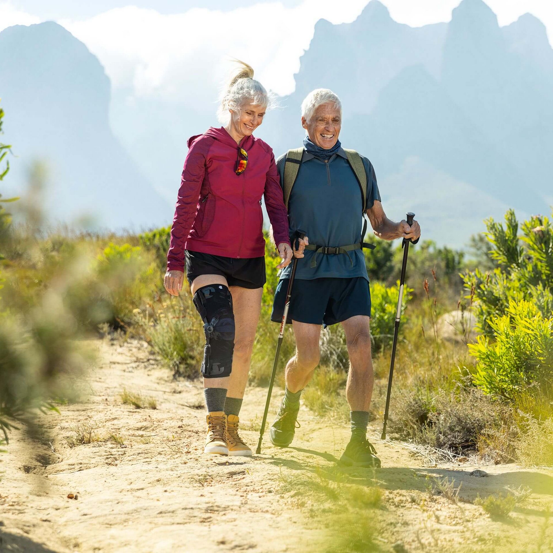  Auf dem Bild sieht man ein älteres Paar, das in einer malerischen Berglandschaft wandert. Die Frau trägt eine GenuTrain OA Orthese, die speziell für Menschen mit Arthrose entwickelt wurde, um ihr Kniegelenk zu unterstützen.