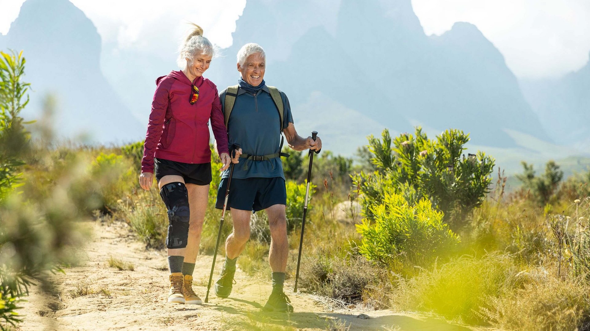  Auf dem Bild sieht man ein älteres Paar, das in einer malerischen Berglandschaft wandert. Die Frau trägt eine GenuTrain OA Orthese, die speziell für Menschen mit Arthrose entwickelt wurde, um ihr Kniegelenk zu unterstützen.