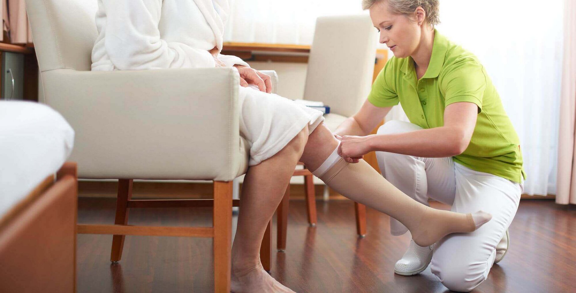 Eine Patientin, welche auf einem Stuhl sitzt, bekommt von einem medizinischen Pflegepersonal Kompressionsstrümpfe von Bauerfeind angezogen.