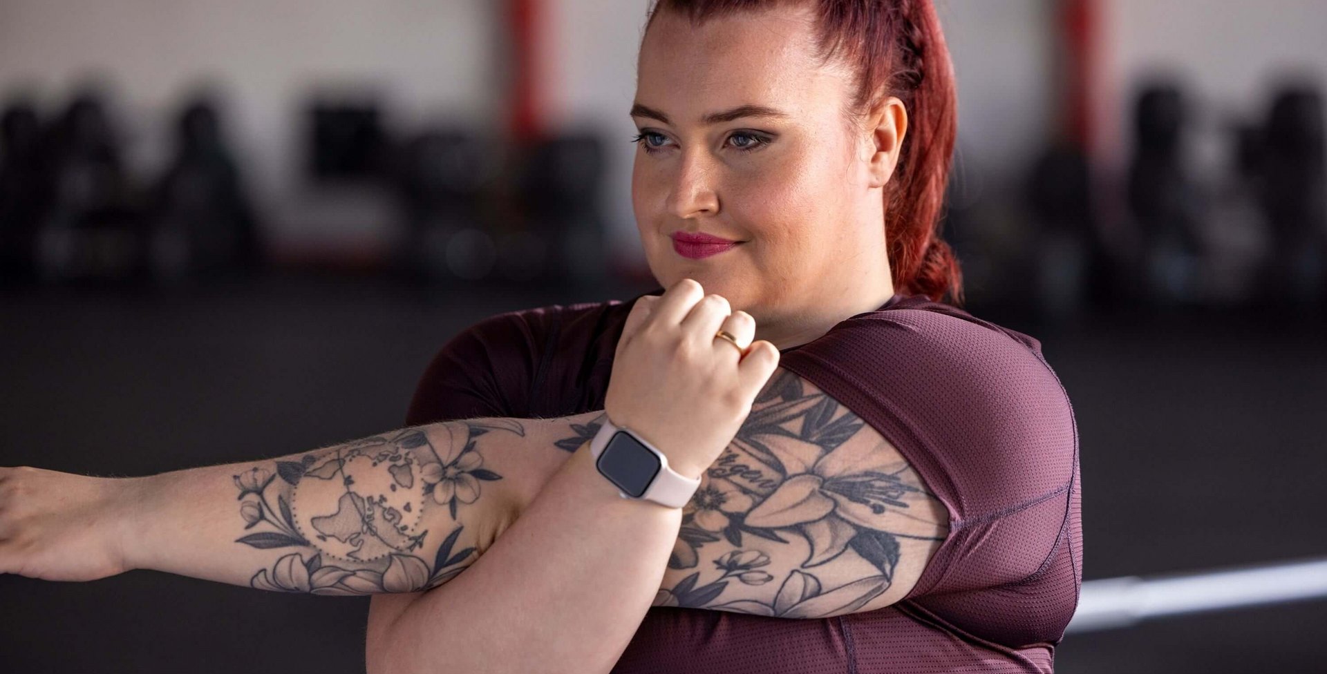 Frau mit roten, geflochtenen Haaren und sichtbaren Tattoos auf beiden Armen dehnt ihren Arm in einem Fitnessstudio. Sie trägt ein bordeauxfarbenes Sportshirt und eine Smartwatch mit hellrosa Armband. Im Hintergrund sind verschwommene Fitnessgeräte zu erkennen.