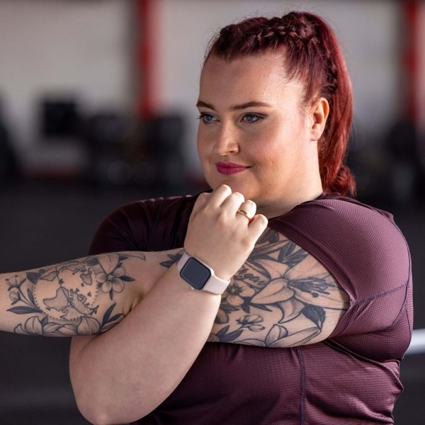 Frau mit roten, geflochtenen Haaren und sichtbaren Tattoos auf beiden Armen dehnt ihren Arm in einem Fitnessstudio. Sie trägt ein bordeauxfarbenes Sportshirt und eine Smartwatch mit hellrosa Armband. Im Hintergrund sind verschwommene Fitnessgeräte zu erkennen.