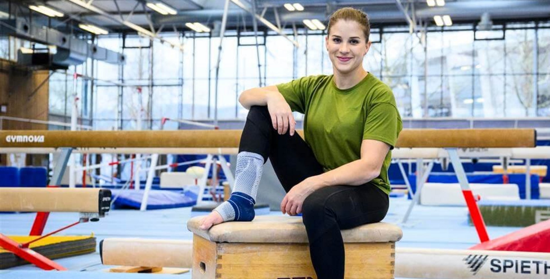Turnerin Elisabeth Seitz sitzt auf einem Holzkasten in einer Sporthalle, trägt ein grünes T-Shirt, schwarze Leggings und die AchilloTrain-Bandage von Bauerfeind am rechten Fuß, umgeben von Turngeräten wie Schwebebalken.