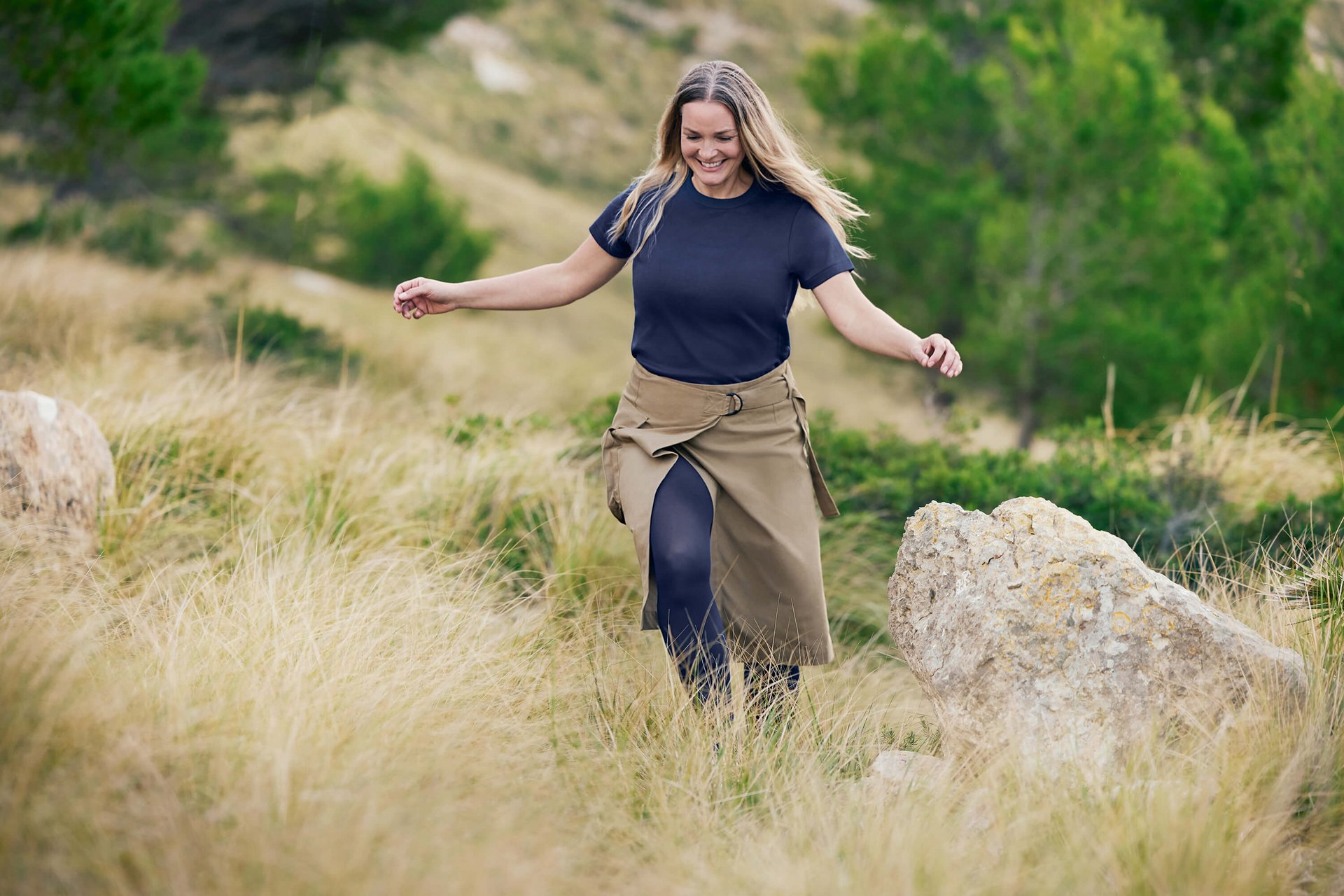 Eine lächelnde Frau mit langen, offenen Haaren geht leichtfüßig durch eine hügelige Naturlandschaft mit hohem Gras und Felsen. Sie trägt ein lässiges dunkelblaues T-Shirt, einen khakifarbenen Wickelrock und VenoTrain Kompressionsstrümpfe in Dunkelblau.