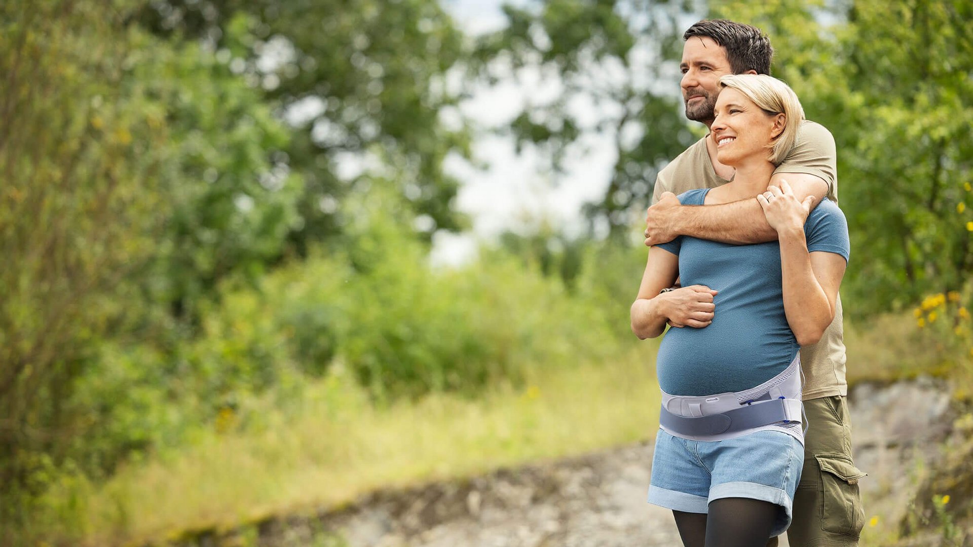Schwangere Frau beim Spaziergang in der Natur, unterstützt durch die Spinova Mum Schwangerschaftsorthese. Ein Mann umarmt sie liebevoll von hinten. 